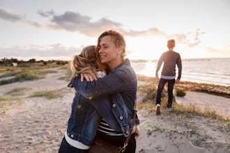 Mor krammer teenage datter på stranden og bror står i baggrunden