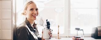 Smilende Kvinde I Solskin Sidder Med Kop Kaffe Foran Et Stort Vindue