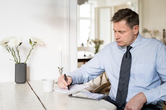 Mand Sidder Og Skriver På Et Stykke Papir Med En Ipad På Bordet